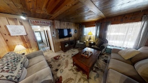 L’intérieur d’un chalet accueillant, avec un plafond en bois, un téléviseur fixé au mur, un canapé, une table basse et un tapis.