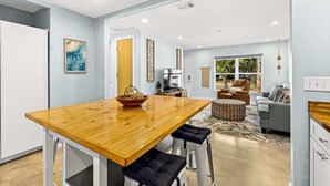 Una cocina moderna con una mesa de madera y taburetes de bar, un área de comedor con un sofá y televisor, y vistas al exterior a través de un ventanal grande.
