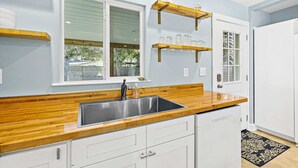 Una cocina con encimera de madera, armarios blancos, un lavamanos de acero inoxidable y una ventana con persianas.