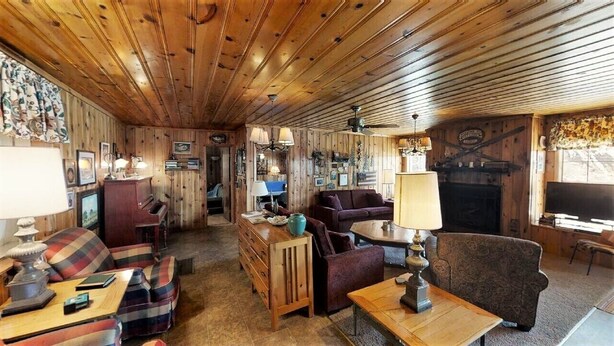 Una habitación acogedora estilo cabaña, con paredes y techo de madera, chimenea, sofá, sillón, mesa y televisión.