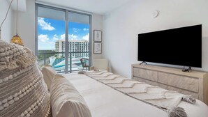A modern living room with a large flat-screen TV, a sofa, a coffee table, and a view of a building through a large window. - One Bedroom Condo King Bed With City Views (Miami)