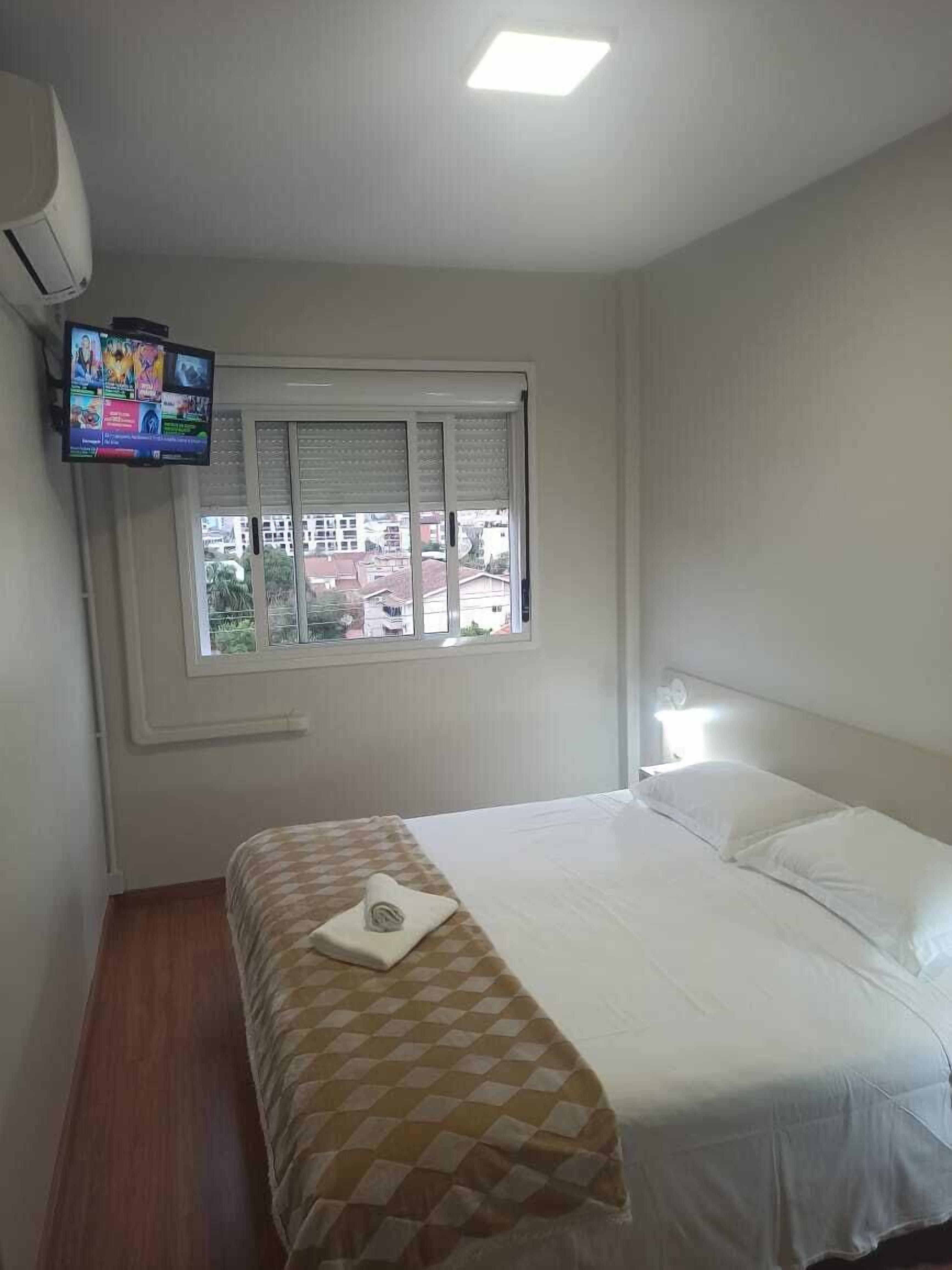 A hotel room with a bed, a television mounted on the wall, a window with blinds, and a view of buildings outside.