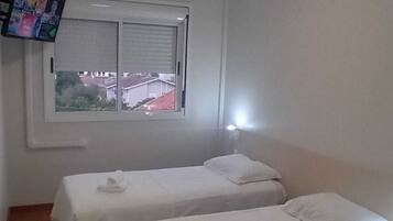 A small, clean room with two single beds, a window with blinds, and a TV mounted on the wall.