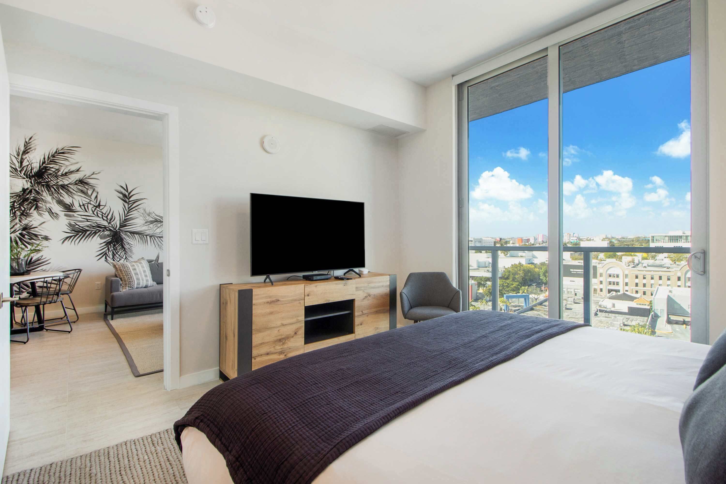 A modern hotel room with a large bed, a flat-screen TV on a wooden console, a city view through large windows, and a dining area with a table and chairs.