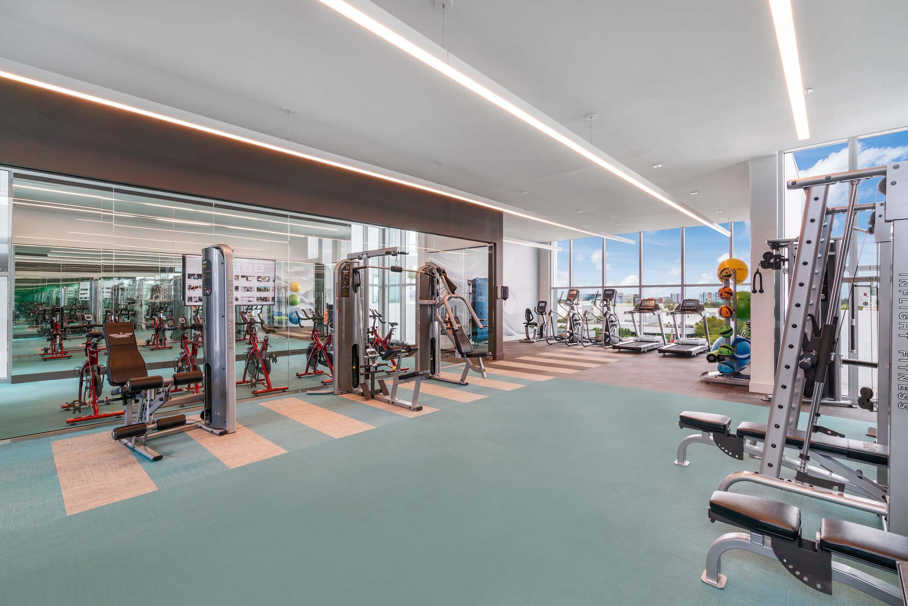 A modern gym with various exercise machines, large windows, and a clean, spacious layout.