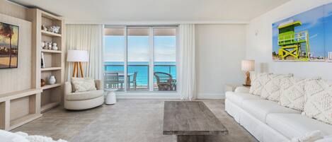 A spacious living room with a large sliding glass door leading to a balcony with a view of the ocean. There is a white sofa, a coffee table, and a bookshelf with decorative items.