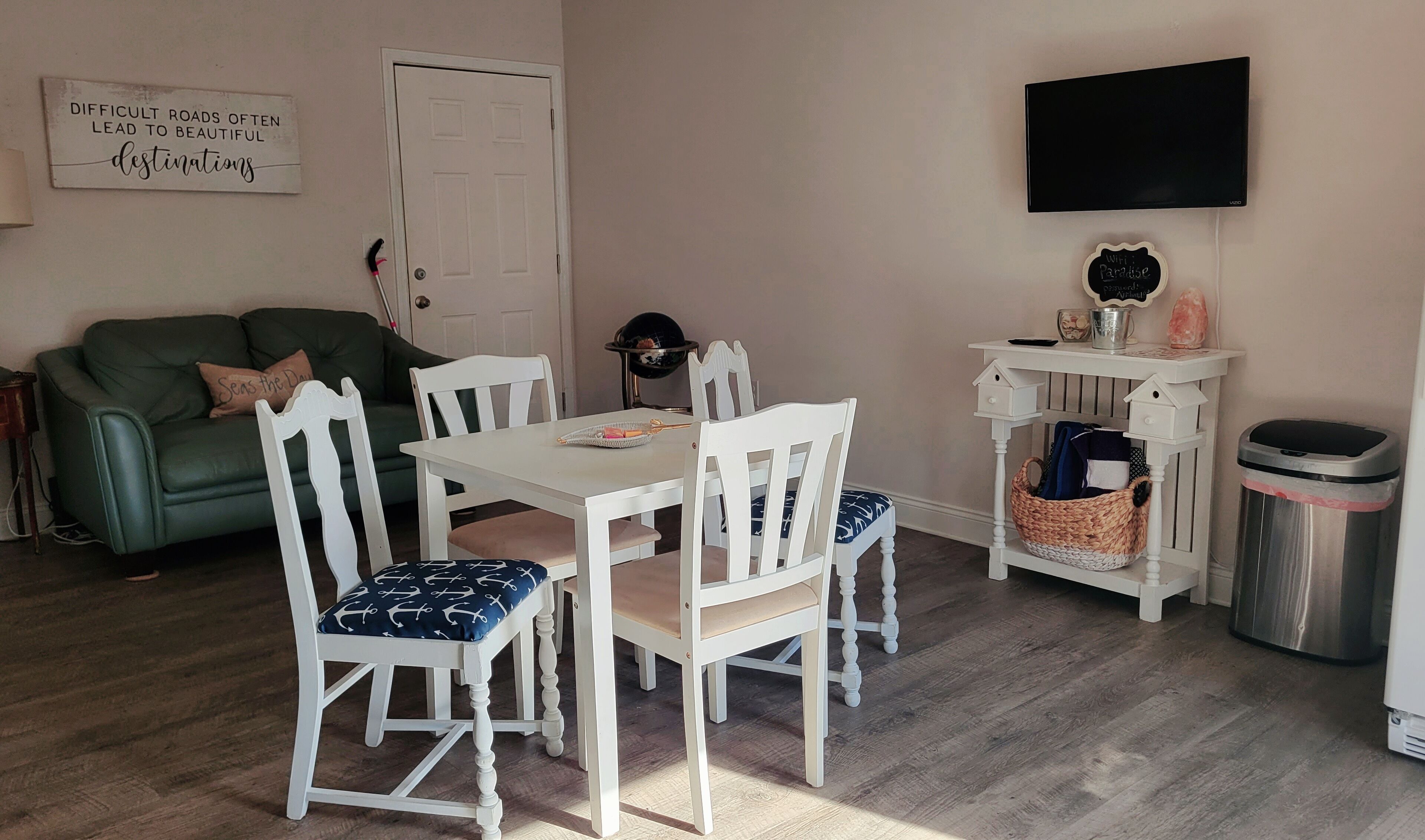 A dining area with a white table and chairs, a green sofa, a TV mounted on the wall, and a trash bin.