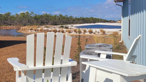 Ein weißer Adirondack-Stuhl und ein kleiner Tisch mit einem Projektor, platziert auf einer Holzterrasse mit Blick auf Strand und Wasser.
