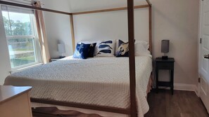 A four-poster bed with a quilted bedspread, a bedside table with a lamp, and a view of a garden through a window.