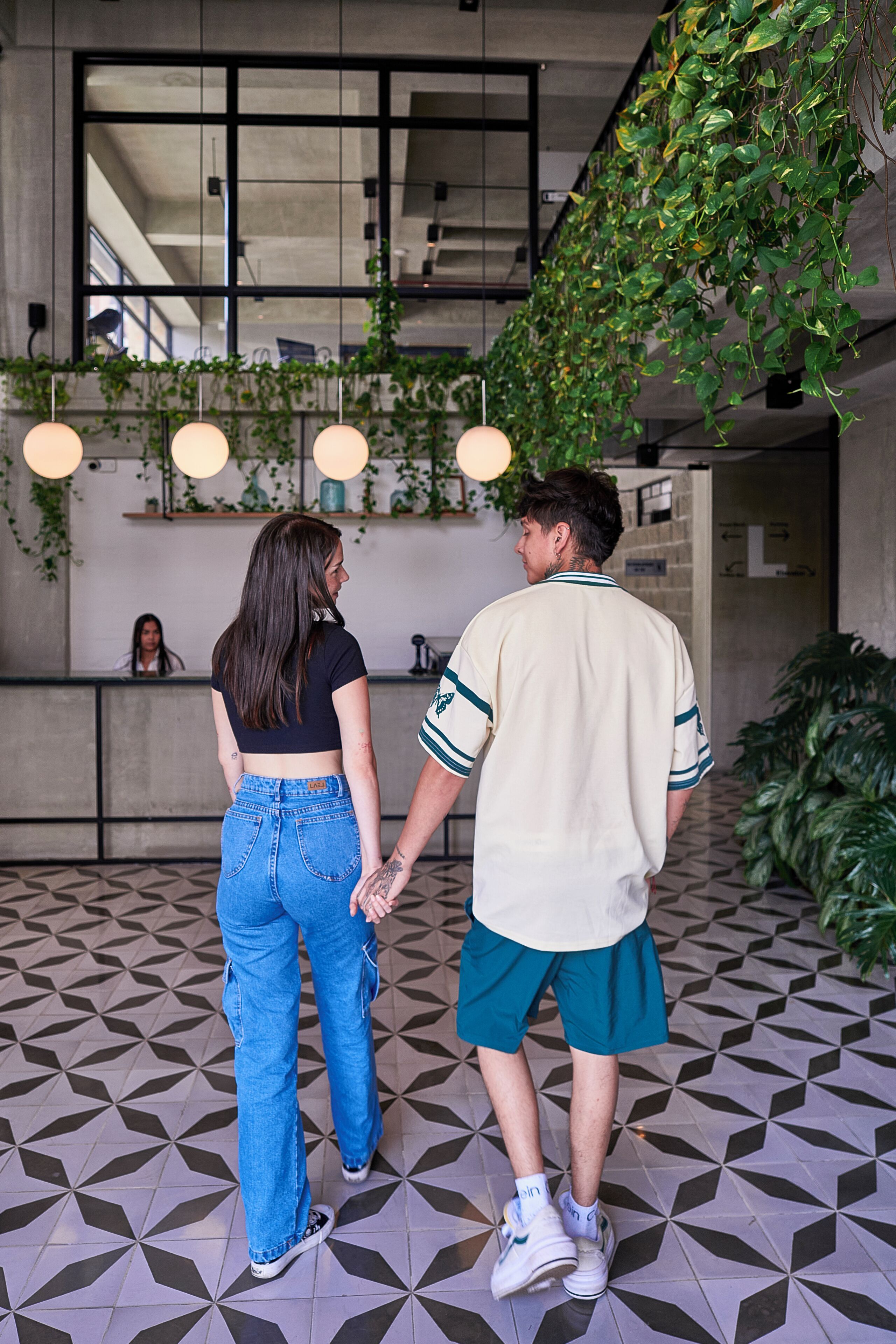 Una pareja tomándose de las manos en un espacio interior moderno con suelo de diseño geométrico, un mostrador de recepción e instalación de plantas colgantes.