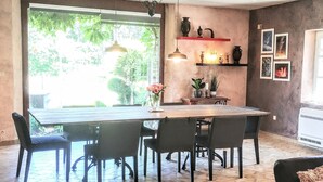 Un comedor con una gran mesa de madera, sillas negras, lámparas colgantes y vistas al exterior con vegetación.