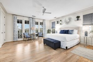 A spacious bedroom with a large bed, a ceiling fan, a small table with chairs, and a view through the large windows.