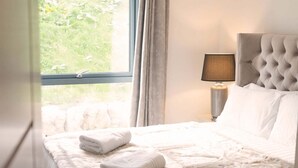 A neatly made bed with a fluffy white blanket and folded towels, a lamp on a bedside table, and a window with curtains. - 8 Loch View Lochtay (Aberfeldy)
