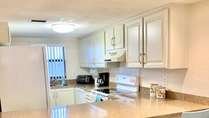 A modern kitchen with white cabinets, a granite countertop, and a built-in oven. There are two bar stools and a window with blinds. - Gorgeous Condo Steps From the Beach! (Daytona Beach Shores)