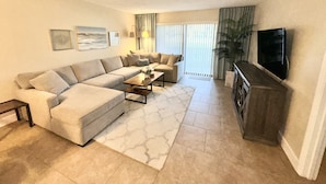 A modern living room with a sectional sofa, a coffee table, and a flat-screen TV mounted on the wall. - Gorgeous Condo Steps From the Beach! (Daytona Beach Shores)