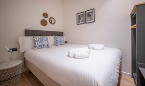 Una cama bien hecha con sábanas blancas y almohadas estampadas en azul, una mesita de noche con lámpara, y una alfombra redonda pequeña.