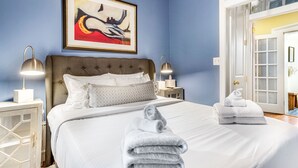 Un lit bien fait, avec du linge de lit blanc et deux serviettes pliées, un tableau abstrait encadré au-dessus du lit, et une porte donnant sur une autre pièce.