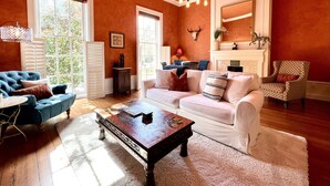 Un salon avec un canapé bleu, un canapé blanc et un fauteuil à motifs, une table basse en bois et une cheminée surmontée d’un miroir.