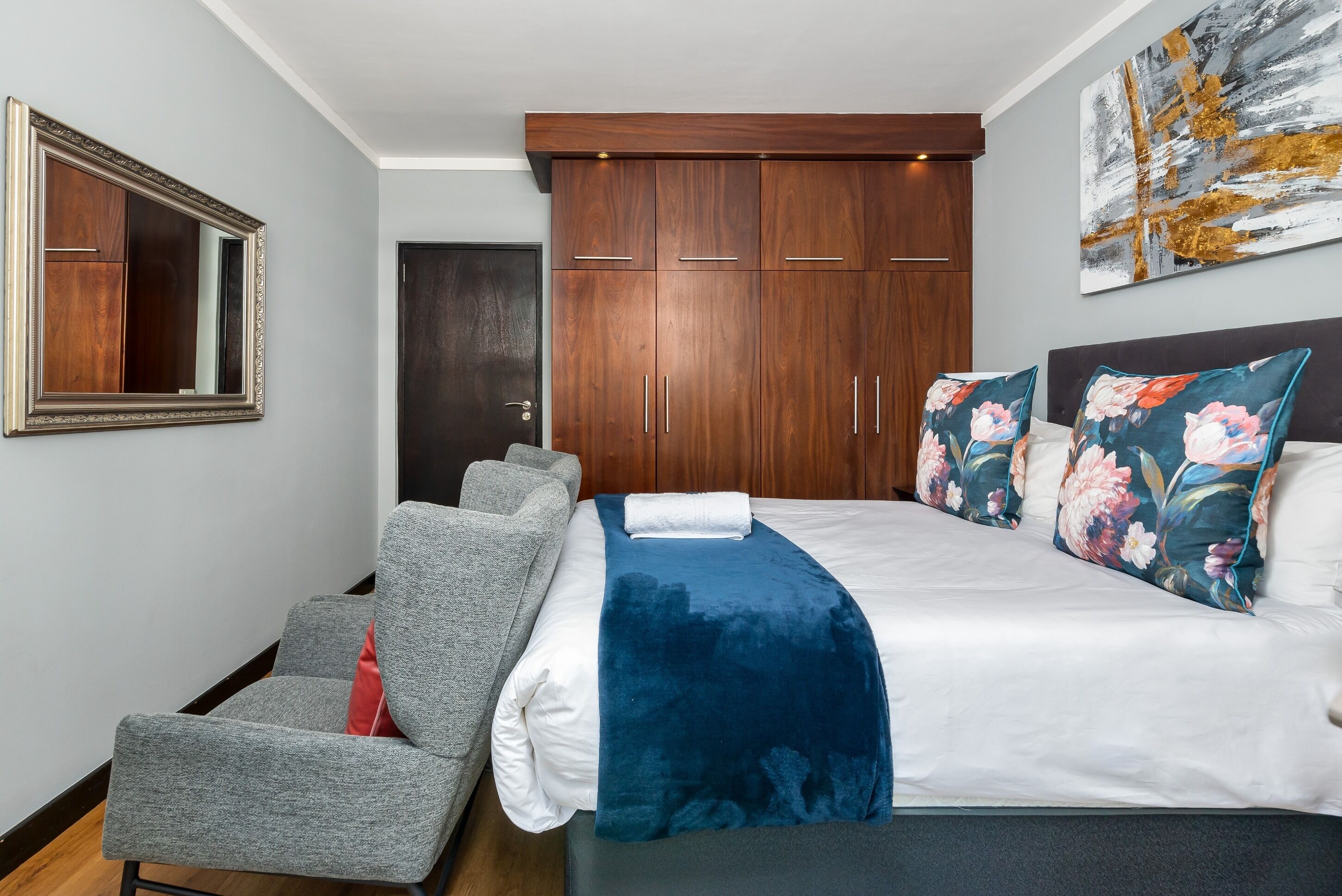 A bedroom with a bed, a grey armchair, a large mirror, wooden wardrobe, and a floral artwork on the wall.