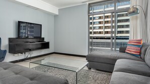 Una sala de estar moderna con un sofá gris, un televisor de pantalla plana montado en la pared, una mesa de centro de vidrio y un amplio ventanal con persianas.