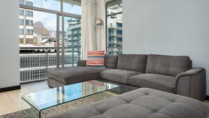 Una sala de estar moderna con un sofá gris, una mesa de centro de vidrio y vistas a un paisaje urbano a través de amplios ventanales.