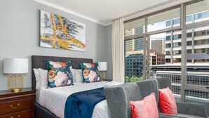 Habitación de hotel moderna con una cama grande, sofá gris y vistas a la ciudad a través de amplios ventanales.