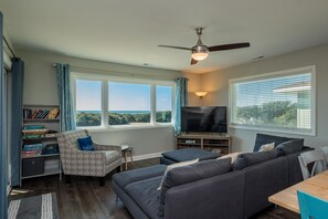 A living room with a sofa, armchair, TV, bookshelf, and large windows.