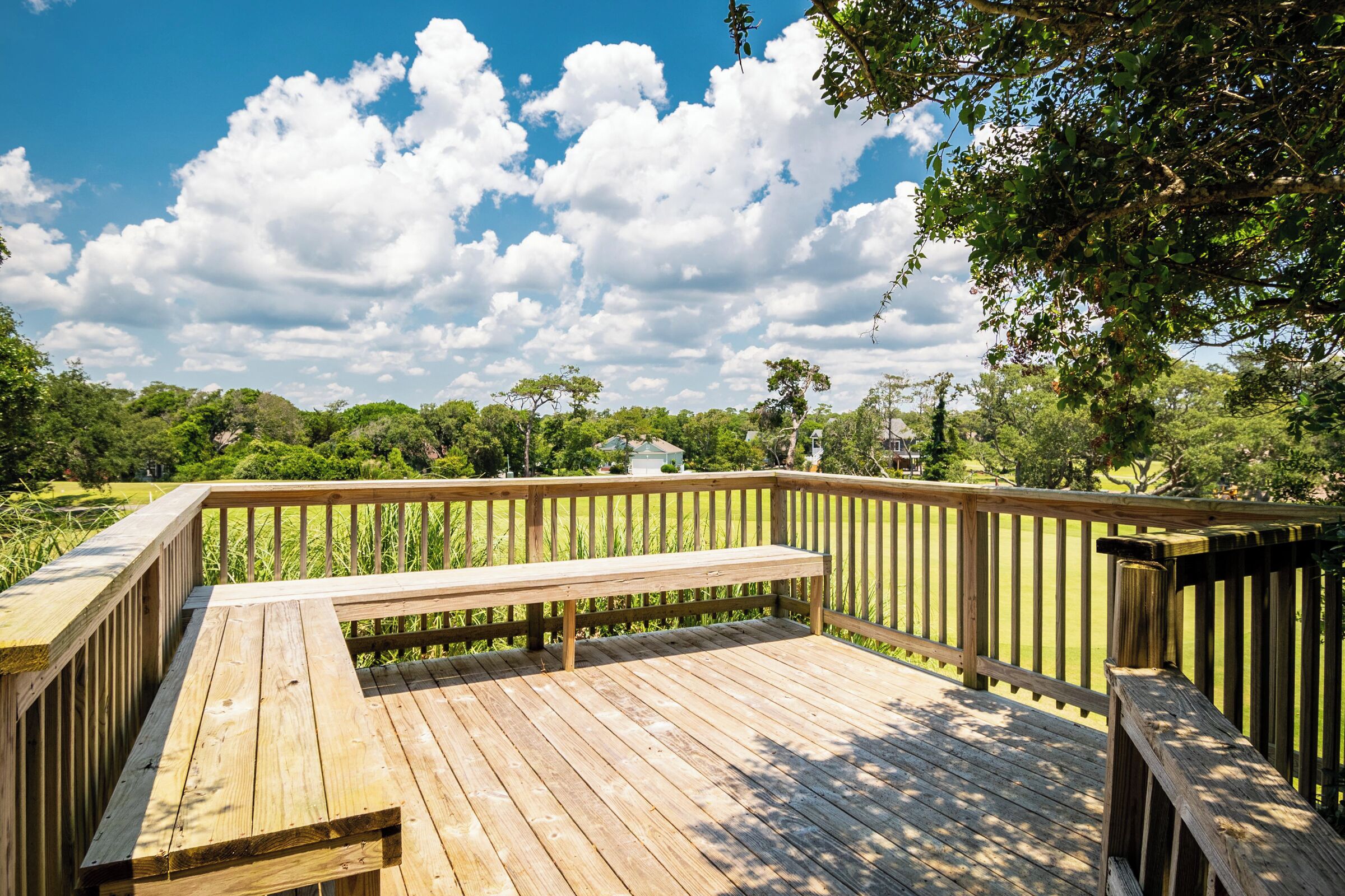 Eine Holzterrasse mit einer Bank, umgeben von üppigem Grün und Bäumen unter einem teilweise bewölkten Himmel.