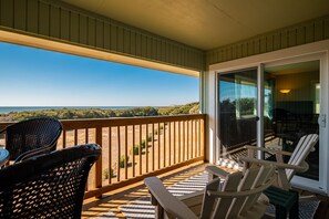 En balkon med fletstole og et bord, med udsigt over en strand og grøn bevoksning.