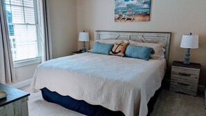 Ein Schlafzimmer mit einem großen Bett, zwei Nachttischen, einem Fenster mit Jalousien und einem Wandbild im Strandhausstil.