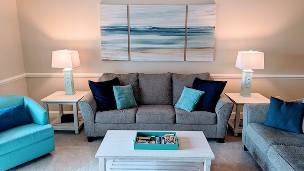 Ein Wohnzimmer mit grauem Sofa, zwei Beistelltischen mit Lampen, einem Couchtisch mit Zeitschriften und einem großen abstrakten Gemälde an der Wand.