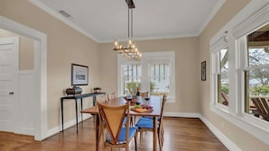A dining area with a wooden table, chairs, and a chandelier. There are windows with white frames and a view of the outdoors.