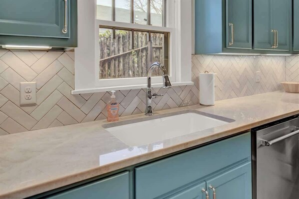 A kitchen with a herringbone tile backsplash, a window with a view of a wooden fence, a stainless steel sink, and teal cabinets.