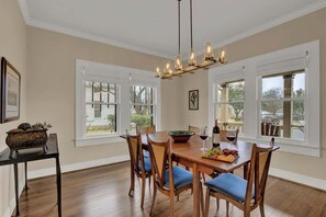 Une salle à manger avec une table en bois, six chaises, un lustre et de grandes fenêtres.