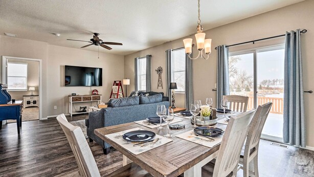 Un coin repas avec une table en bois dressée pour quatre personnes, un ventilateur de plafond et une grande fenêtre avec des rideaux.