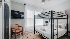 A bunk bed room with a ceiling fan, a TV mounted on the wall, a chair, and a window with sheer curtains.