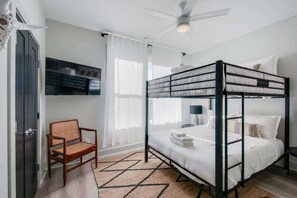 A bunk bed room with a ceiling fan, a TV mounted on the wall, a chair, and a window with sheer curtains.