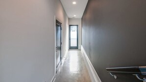 A narrow hallway with a door at the end, a light fixture on the ceiling, and a metal handrail on the right wall.