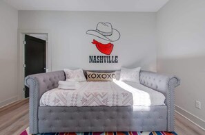 Un dormitorio con una cama tapizada gris, un adorno de pared con un sombrero de vaquero y una bandana roja con el texto 