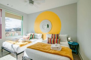 Habitación de hotel con dos camas, una pared grande de color amarillo, un espejo redondo y una mesita de noche pequeña con una lámpara.