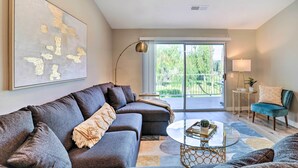 A modern living room with a sectional sofa, a glass coffee table, and a view of the outdoors through a sliding glass door. - Chic Palm Desert Condo w/ Resort-Style Amenities! (Palm Desert)