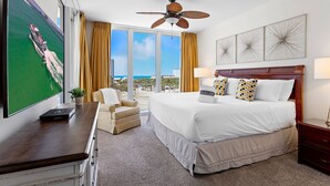 A hotel room with a large bed, a desk, a chair, a ceiling fan, and a TV displaying a beach scene. - Silver Shells St Lucia 501 - Beachfront, Heated Community Pool + Hot Tub! (Destin)