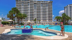 A hotel pool area with a large building in the background, palm trees, and people swimming and relaxing. - Silver Shells St Lucia 501 - Beachfront, Heated Community Pool + Hot Tub! (Destin)