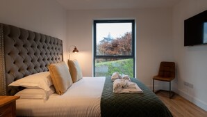 Une chambre moderne avec une tête de lit capitonnée, une grande fenêtre donnant sur les arbres, un téléviseur à écran plat et un fauteuil.