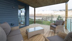 Un espace extérieur abrité doté d'une terrasse en bois, de meubles en osier et d'une porte en verre donnant sur une pièce entièrement vitrée.