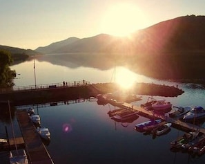 A marina with boats docked, a pier extending into calm water, and mountains in the background.