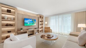 A modern living room with a flat-screen TV, white sofas, a wooden coffee table, and a wicker chair.