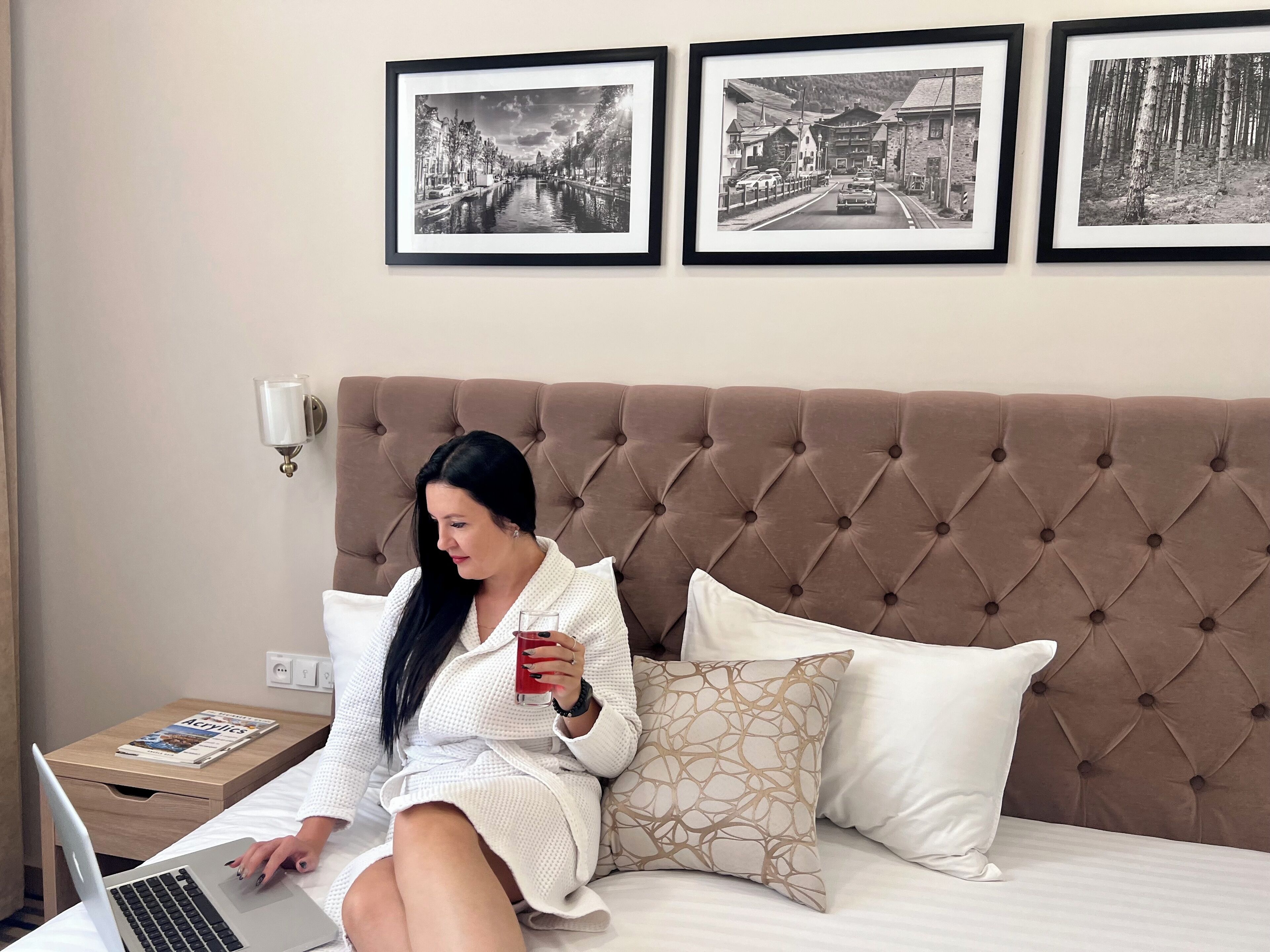 A woman sitting on a bed with a laptop, holding a glass of red drink, with three framed pictures on the wall behind her.
