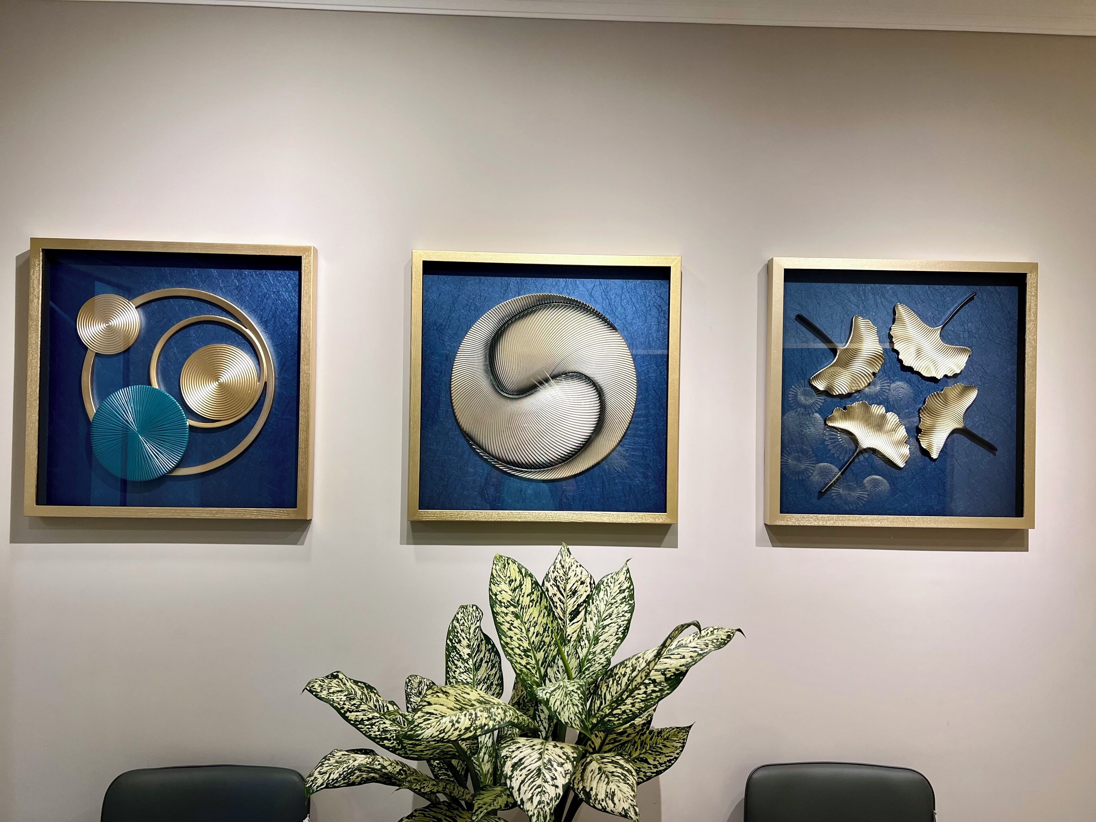 Three framed abstract art pieces on a wall, a potted plant in the foreground, and two chairs.
