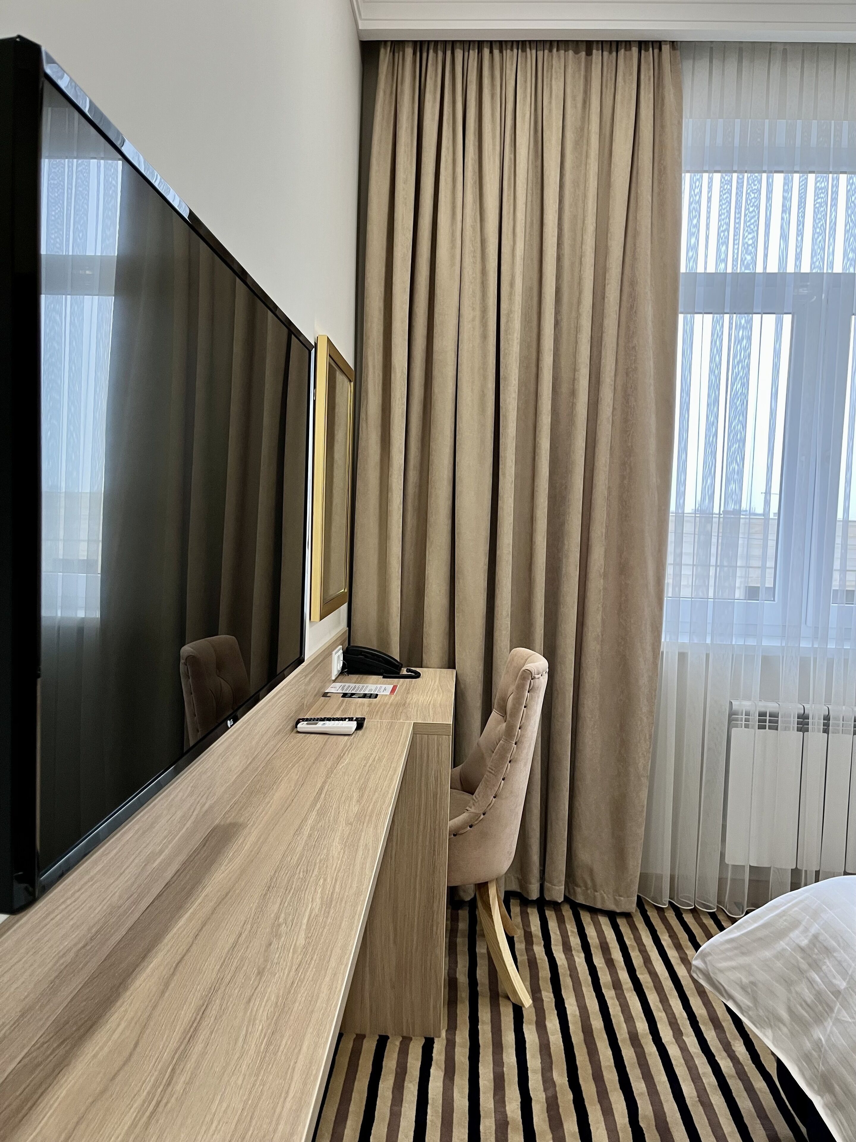 Een hotelkamer met een grote flatscreen televisie, een houten bureau met telefoon en afstandsbediening, een beige fauteuil en een bed met een gestreept tapijt.